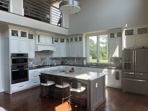 A modern kitchen with white cabinetry, a central island with bar stools, a stainless steel refrigerator, and a view of the outdoors through large windows.
