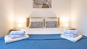A neatly made bed with a blue quilt, two patterned pillows, and folded towels on either side. - A house surrounded by forest. (Valtura)