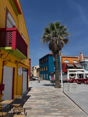 A street with colorful buildings, a palm tree, and outdoor seating. - Our rental in Barcarès for 4 people. Pool, AC, beach, and Lydia within walking distance (Le Barcares)