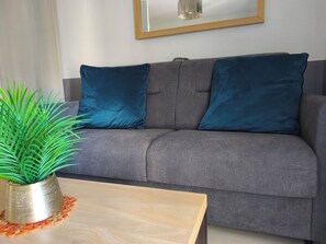 A grey sofa with two dark blue cushions, a wooden coffee table with a plant, and a framed mirror on the wall.