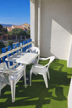 A balcony with a white table and chairs, artificial grass, and a view of buildings in the background. - Our rental in Barcarès for 4 people. Pool, AC, beach, and Lydia within walking distance (Le Barcares)