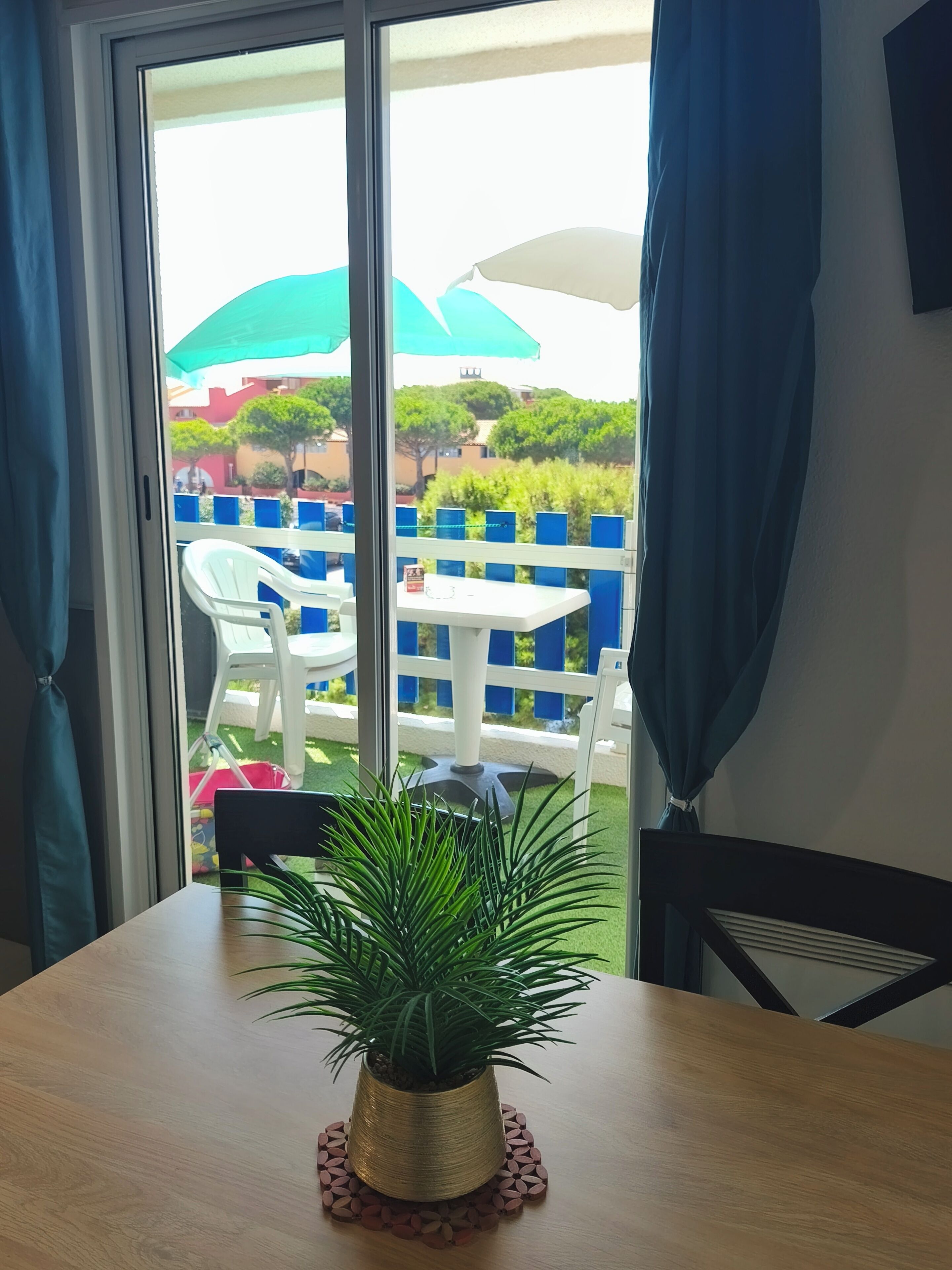 A room with a view of an outdoor area featuring a white table, a potted plant, and a blue curtain.