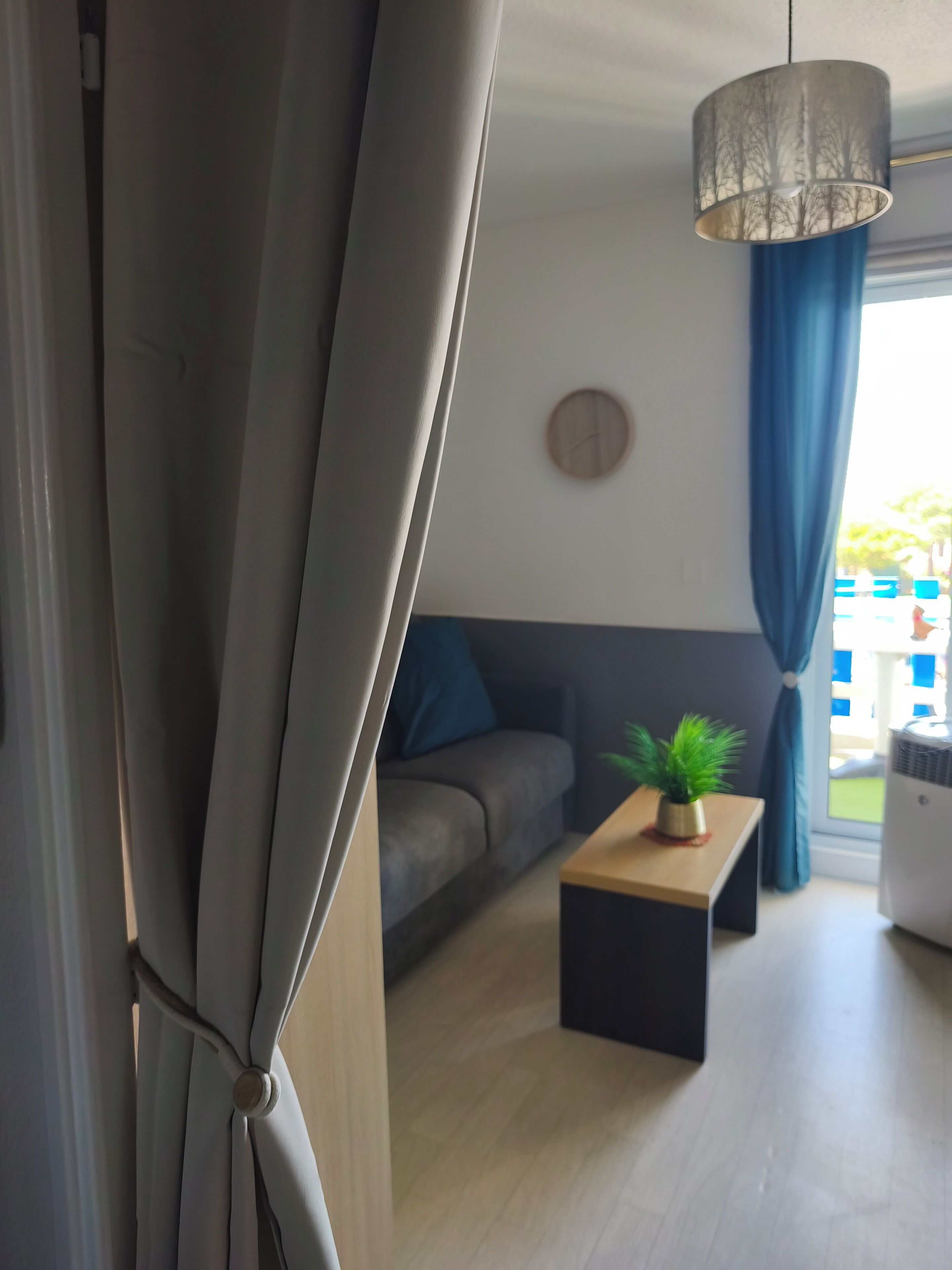 A modern living room with a sofa, a coffee table with a plant, a hanging light fixture, and a window with curtains.