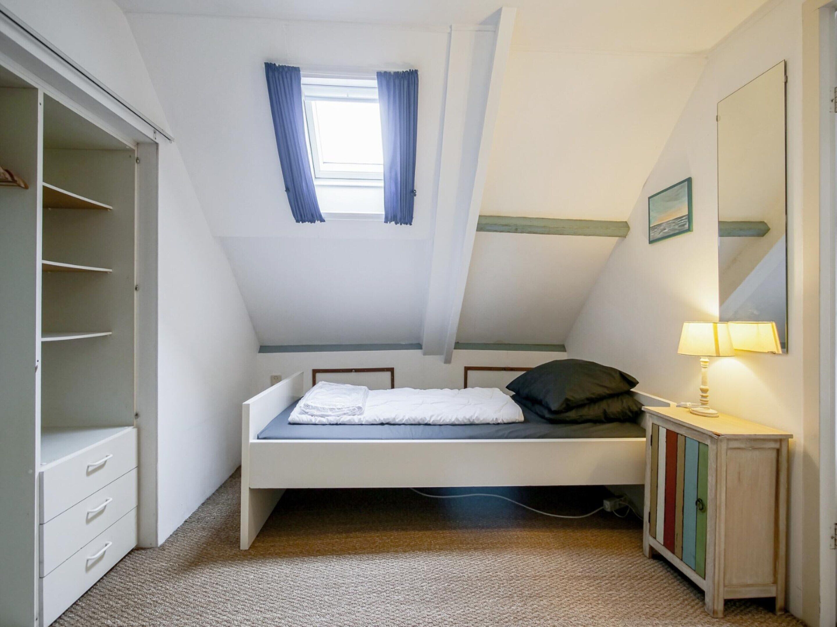 A bedroom with a sloped ceiling, a bed, a wardrobe, a bedside table with a lamp, and a window with curtains.