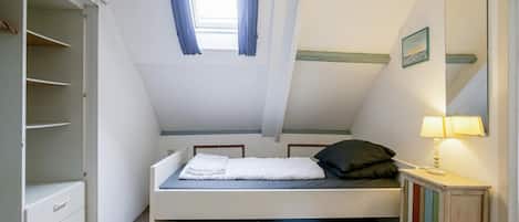 A bedroom with a sloped ceiling, a bed, a wardrobe, a bedside table with a lamp, and a window with curtains.