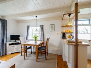 Quarto com mesa de jantar de madeira, cadeiras, televisão, janela com cortinas, área de cozinha com prateleiras e luminária pendente.