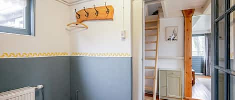A room with a radiator, a wooden floor, a window with a curtain, a wooden shelf with hooks, and a door leading to another room.