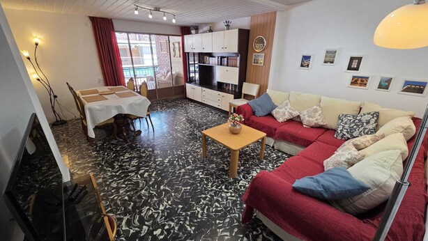 En stue med sofa, sofabord, spisebord med stole, fjernsyn og et køkkenområde.