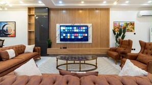 A modern living room with a large flat-screen TV, brown sofas, a glass coffee table, and wooden paneling on the wall. - Beautiful Waterfront 4-bedroom Terrace Duplex in vibrant Lekki (Lekki)