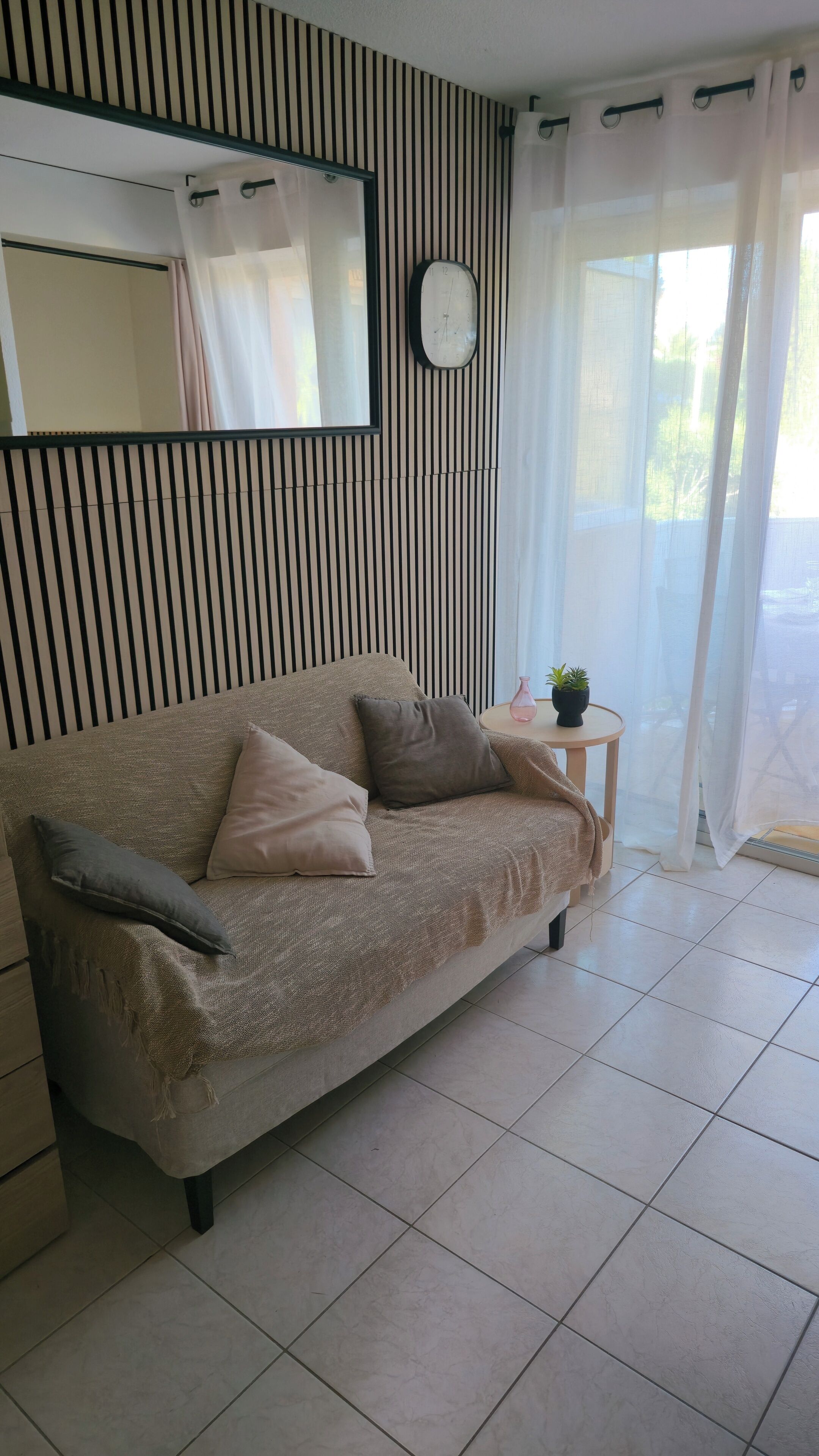Een woonkamer met een bank, een bijzettafeltje met een plant, een klok aan de muur en een raam met gordijnen.