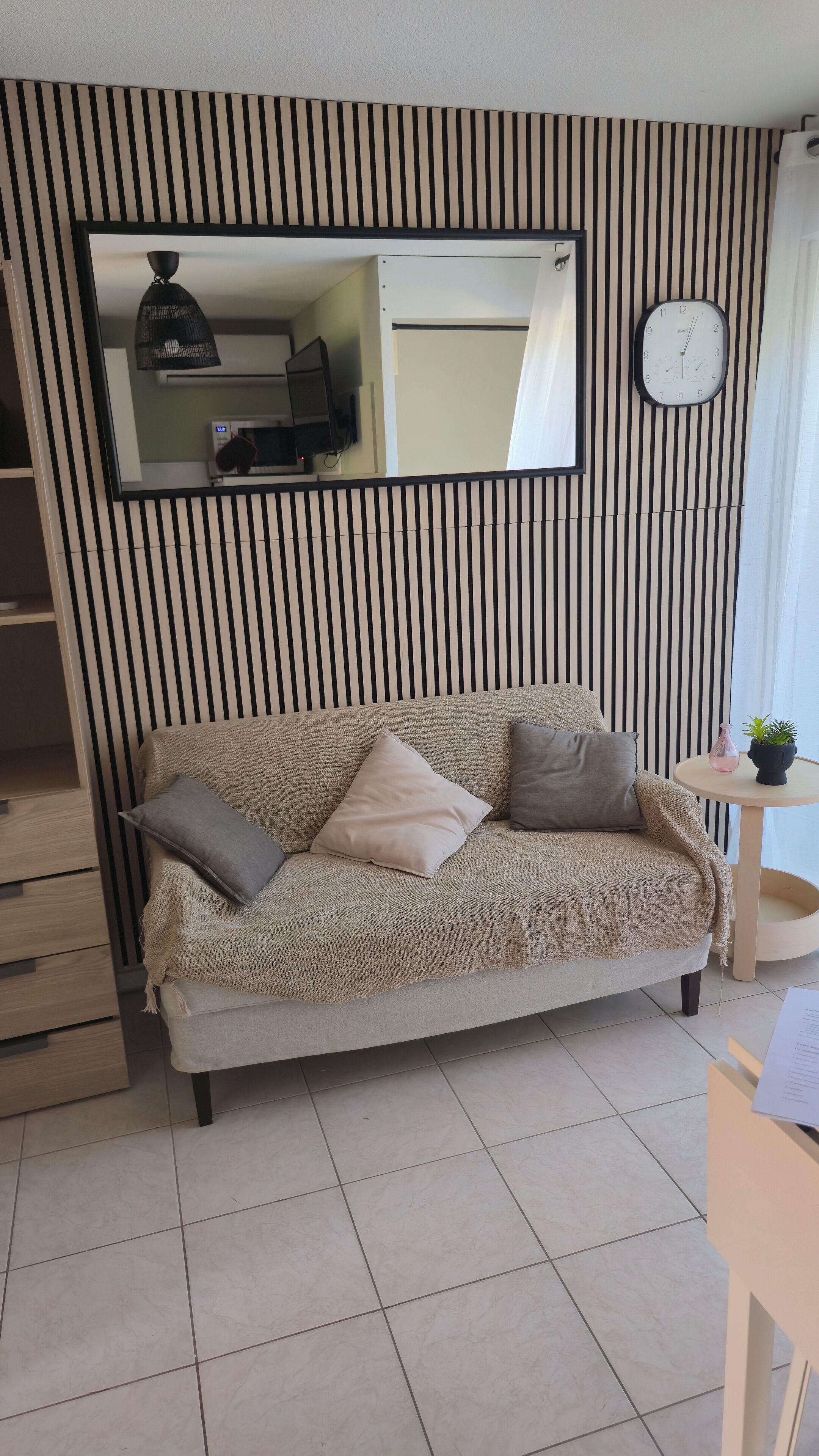 Un salon moderne avec un canapé, des coussins, une table ronde ornée d’une plante, une horloge et un miroir accroché au mur.