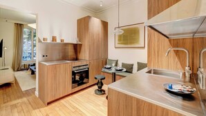 A modern kitchen with wooden cabinets, a built-in oven, a dining area with a round table, and a large window. - Cozy Appartement a/c 1br/2p- Montmartre (Paris)