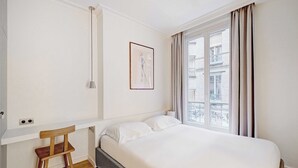 A bedroom with a bed, a chair, a window with curtains, a framed picture on the wall, and a light fixture. - Cozy Appartement a/c 1br/2p- Montmartre (Paris)