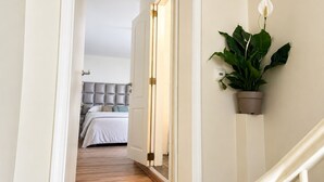 Ein Flur, der in ein Schlafzimmer mit einem Bett, einer Topfpflanze, einer Wandleuchte und einem Thermostat führt.