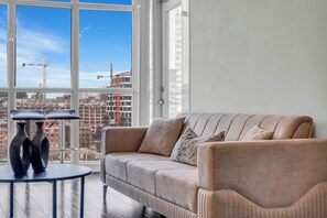 A modern living room with a beige sofa, a round table with two vases, and a large window offering a city view with construction cranes. - The Perfect Downtown Stay Hotel|Steps to Jays, CN Tower & Waterfront W/Parking (Toronto)