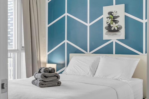 A neatly made bed with white linens, a stack of folded towels, and a wall with a geometric pattern.