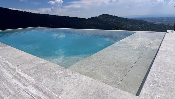 En rektangulär infinitypool med en tydlig, blå kant, omgiven av ett ljust grått, kaklat däck, och med en bergig landskap och en delvis molnig himmel som bakgrund.