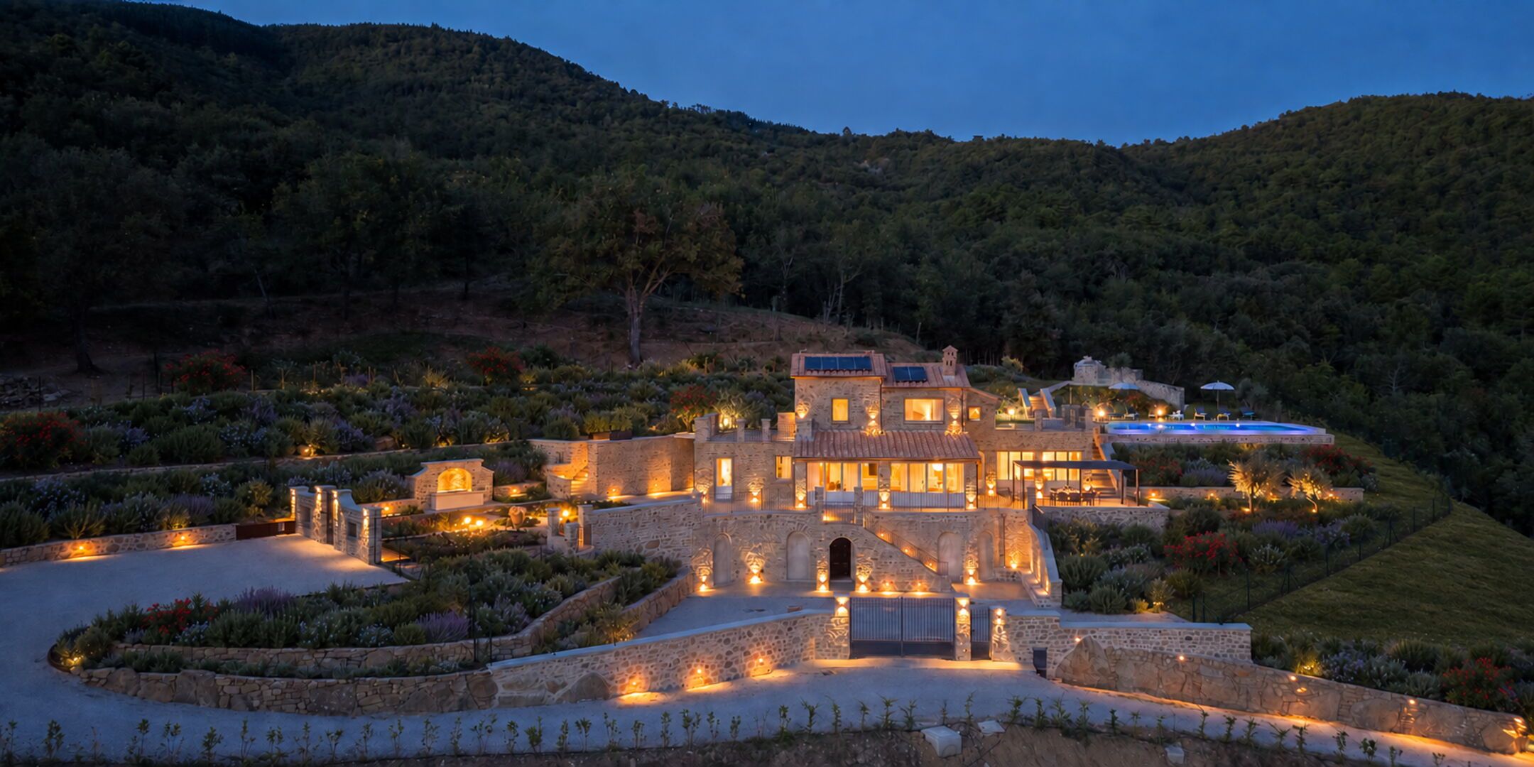 Un grand bâtiment en pierre, bien éclairé, à plusieurs niveaux, doté d’une piscine et entouré de jardins aménagés et de collines.