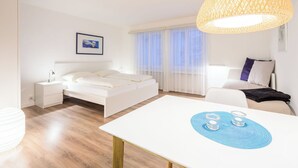 Ein modernes Hotelzimmer mit einem Bett, Nachttisch, Schreibtisch mit Stuhl und einem großen Fenster mit leichtem Vorhang.