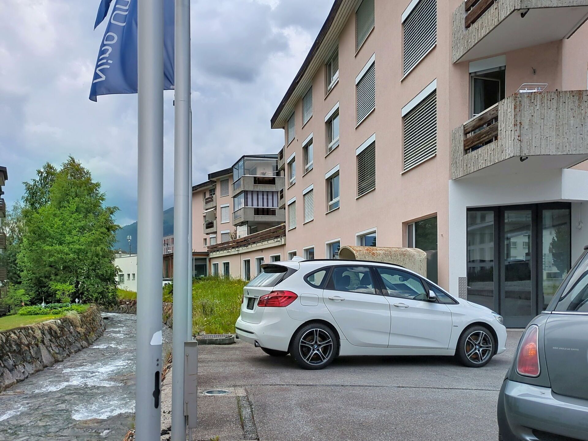 Un immeuble d’habitation doté de nombreux balcons, une voiture blanche garée et un mât avec un drapeau.