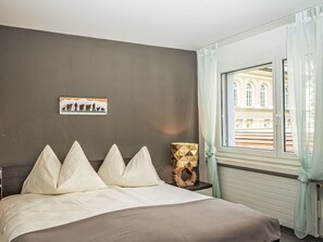 Ein Schlafzimmer mit Bett, Nachttisch, Lampe, einem Fenster mit Vorhängen und einem Wandbild.