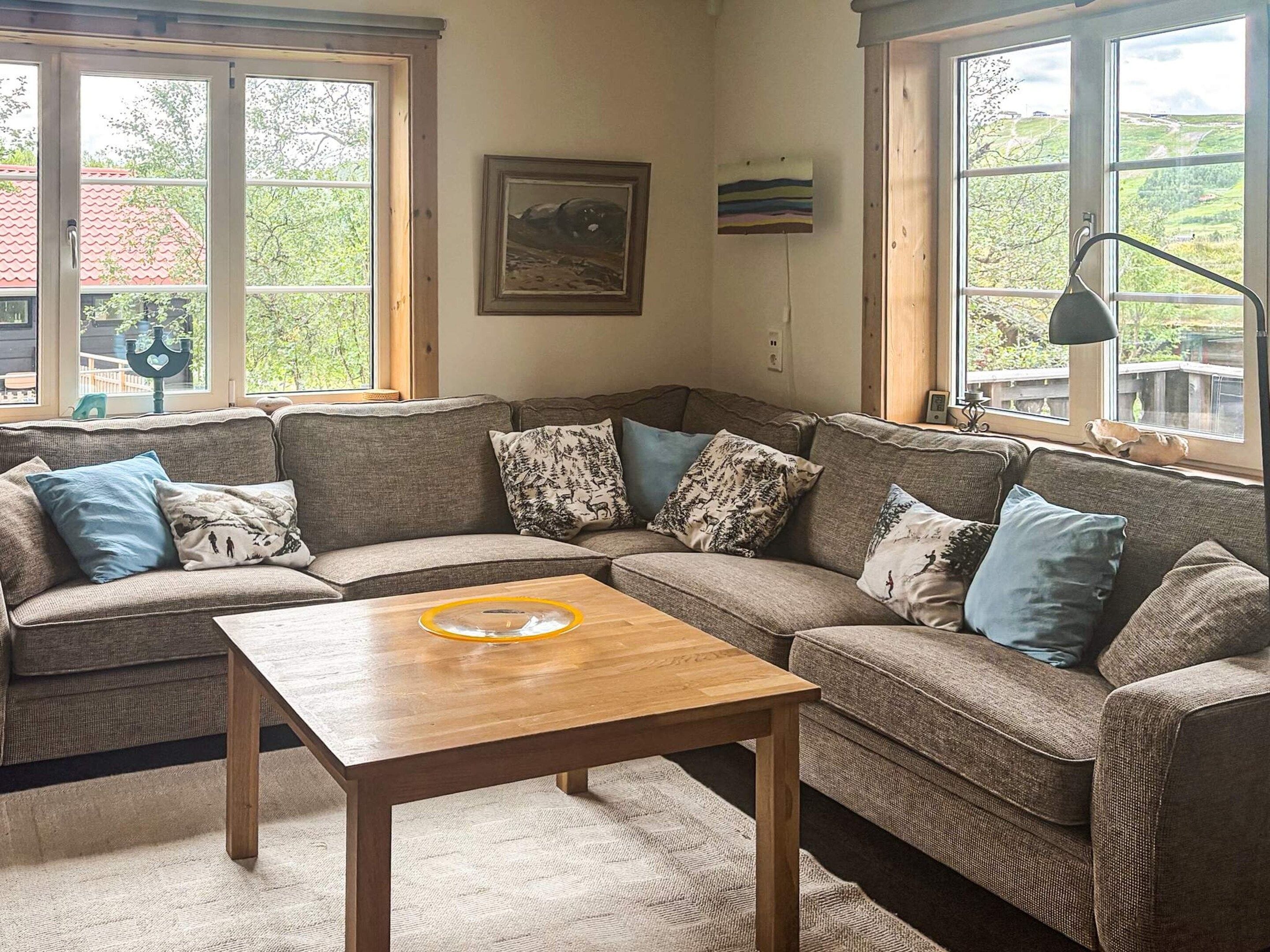 Una sala de estar acogedora con un sofá modular, una mesa de centro de madera y ventanas amplias con vistas a zonas verdes.