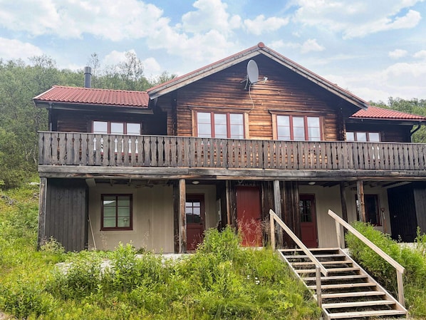 A two-story wooden house with a balcony, a satellite dish, and a staircase leading to the entrance.