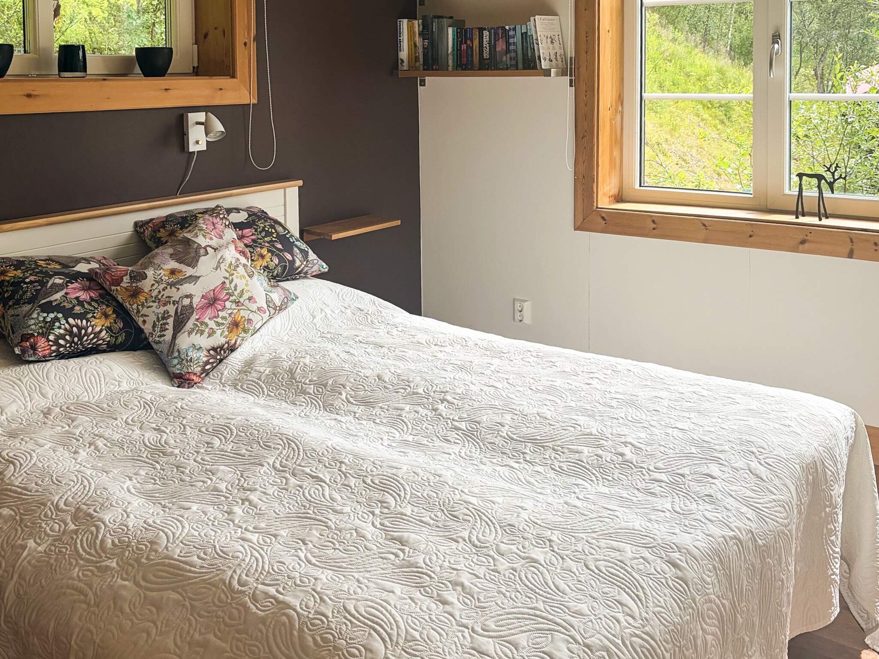 Een slaapkamer met een bed, kussens, een nachtkastje, een raam en een boekenkast.