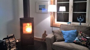 Ett mysigt vardagsrum med en vedkamin, en soffa med kuddar, en lampa och ett fönster med utsikt.