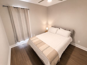 Ein Schlafzimmer mit einem Bett, einem Nachttisch, einer Lampe, einem Fenster mit Vorhängen und einem Deckenventilator.