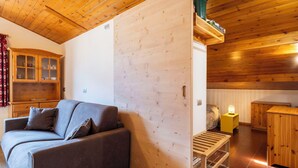 Ein gemütliches Zimmer mit Holzbalkendecke, einem grauen Sofa, einem Holzschrank und einem kleinen Tisch mit Lampe.