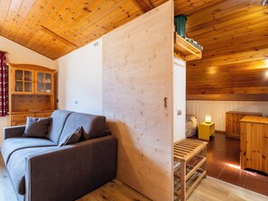 Quarto aconchegante com teto de madeira, sofá cinza, armário de madeira e pequena mesa com abajur.