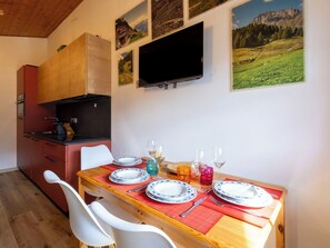 Uma sala de jantar com uma mesa de madeira posta para dois, uma televisão presa na parede e uma cozinha com um armário vermelho.