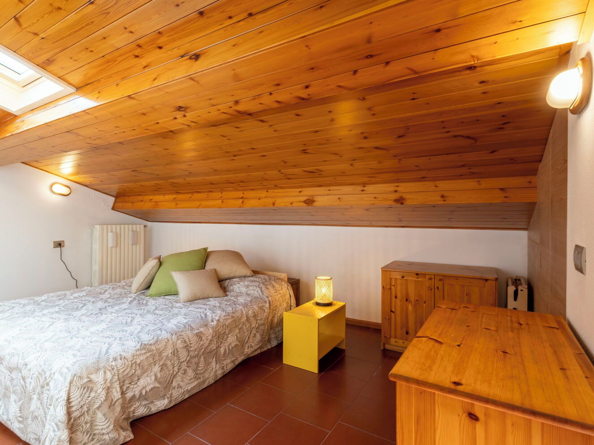 A bedroom with a wooden ceiling, a bed with patterned bedding, a bedside lamp, and a wooden cabinet.