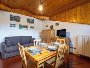 Ein gemütliches Zimmer mit Holzbalkendecke, einem grauen Sofa, einem Esstisch mit Tellern und Gläsern sowie einem Fernseher auf einer Holzkommode.