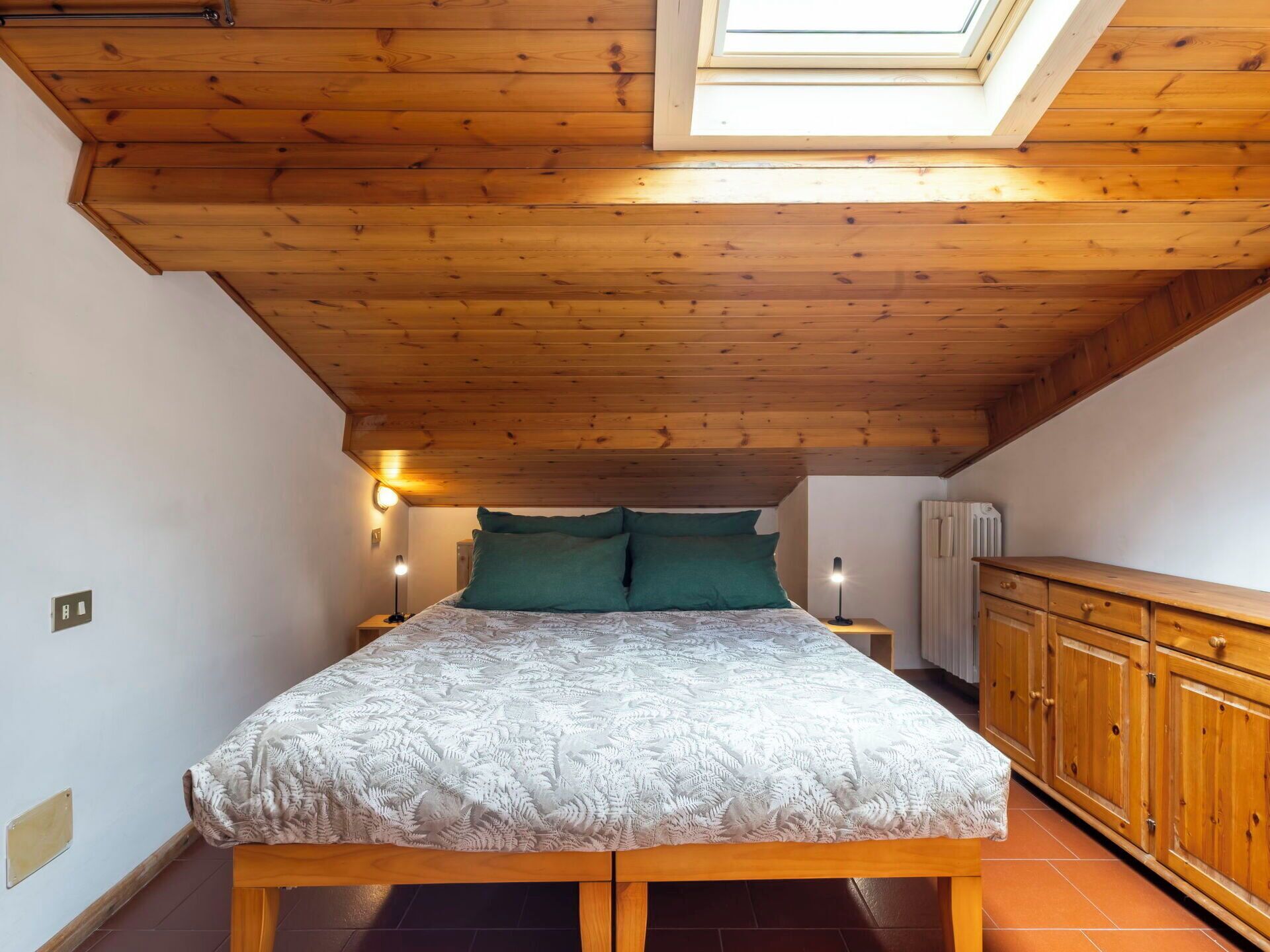 A bedroom with a wooden ceiling, a skylight, a bed with green pillows, bedside lamps, and a wooden cabinet.