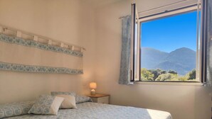Camera da letto con un letto, un comodino con una lampada, una finestra vista montagne e tende a motivi.