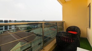 Un balcon doté d’une balustrade en verre, deux fauteuils en osier avec des coussins rouges, et une vue sur les immebles.