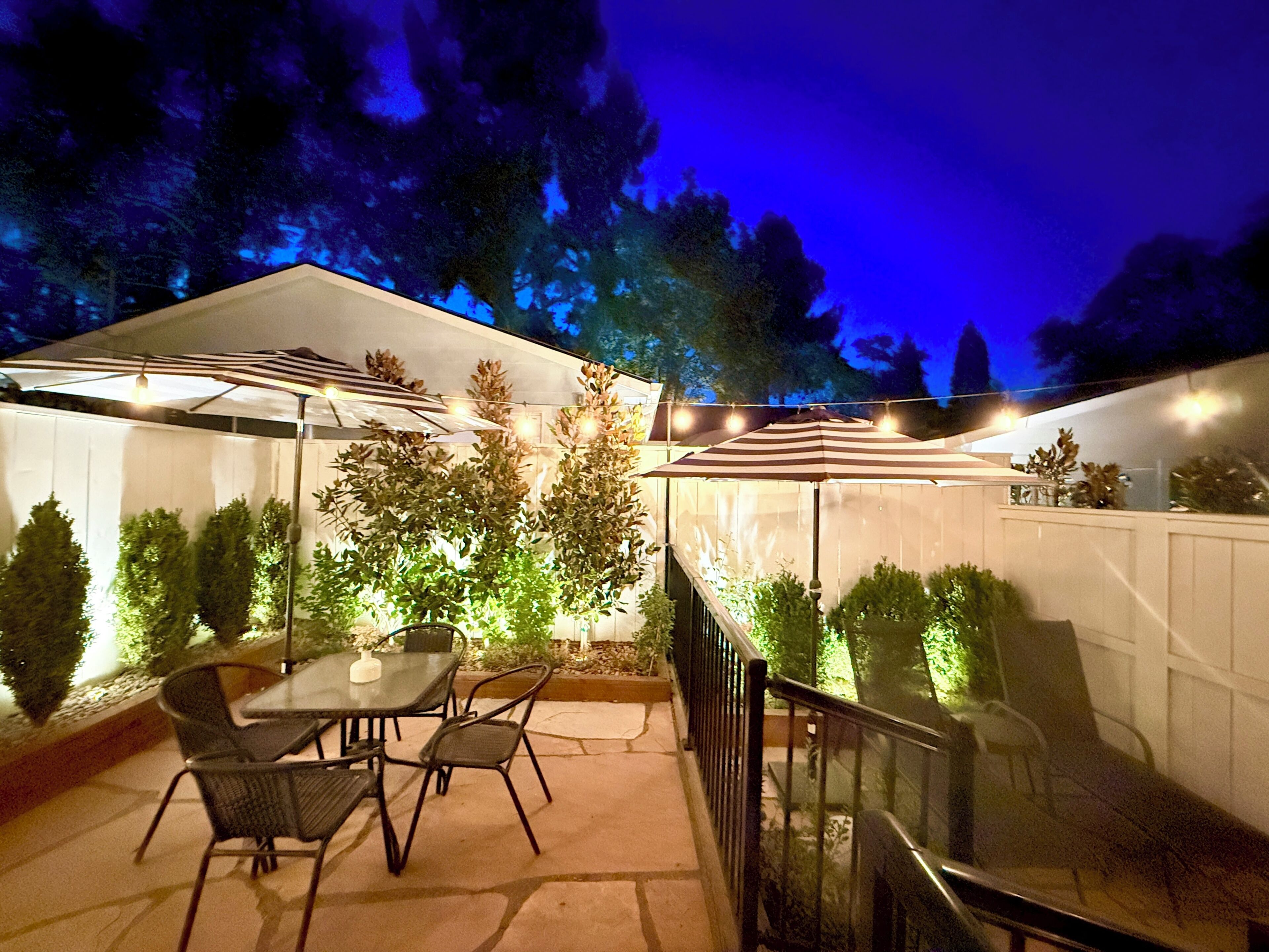 Un espace extérieur aménagé en patio, avec des tables et des chaises, entouré de plantes et d’une clôture blanche.