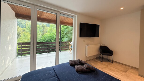 A bedroom with a bed, a TV, a chair, and a view of greenery through a sliding door.