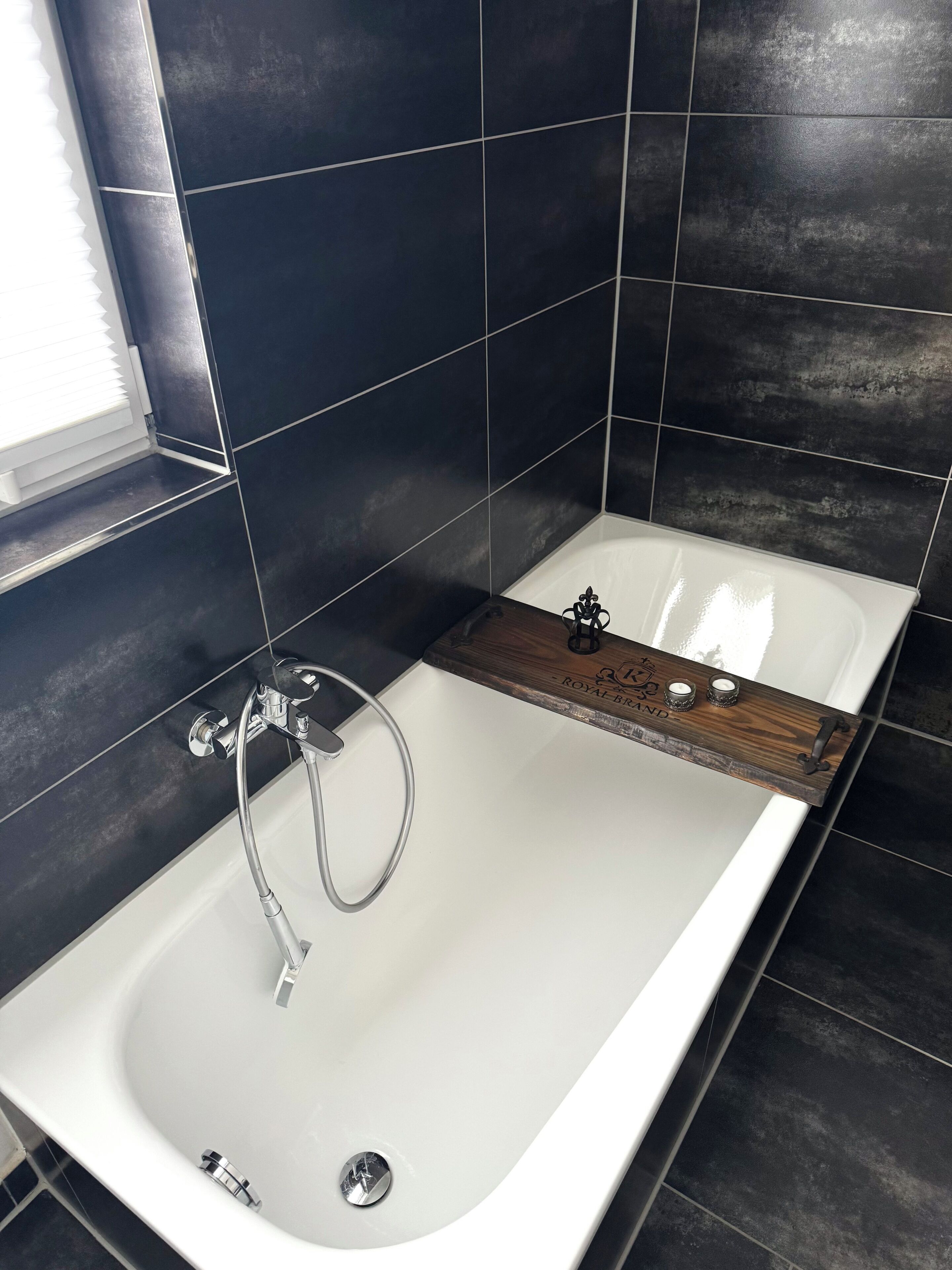 Un baño moderno con una bañera blanca, paredes de azulejos oscuros y una bandeja de madera con velas.