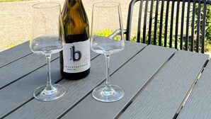 A bottle of wine and two glasses on a table outdoors with a chair and greenery in the background.