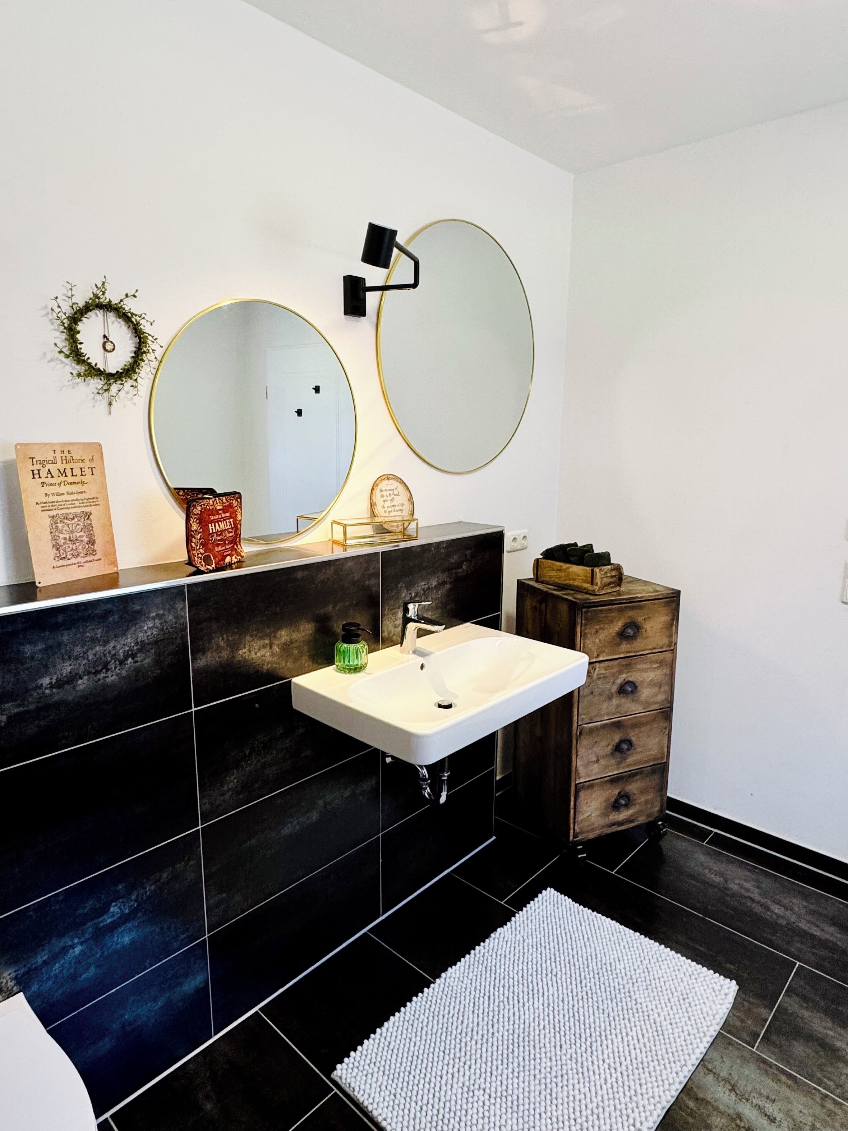 A modern bathroom with a white sink, two round mirrors, a wooden dresser, and a black tiled floor.