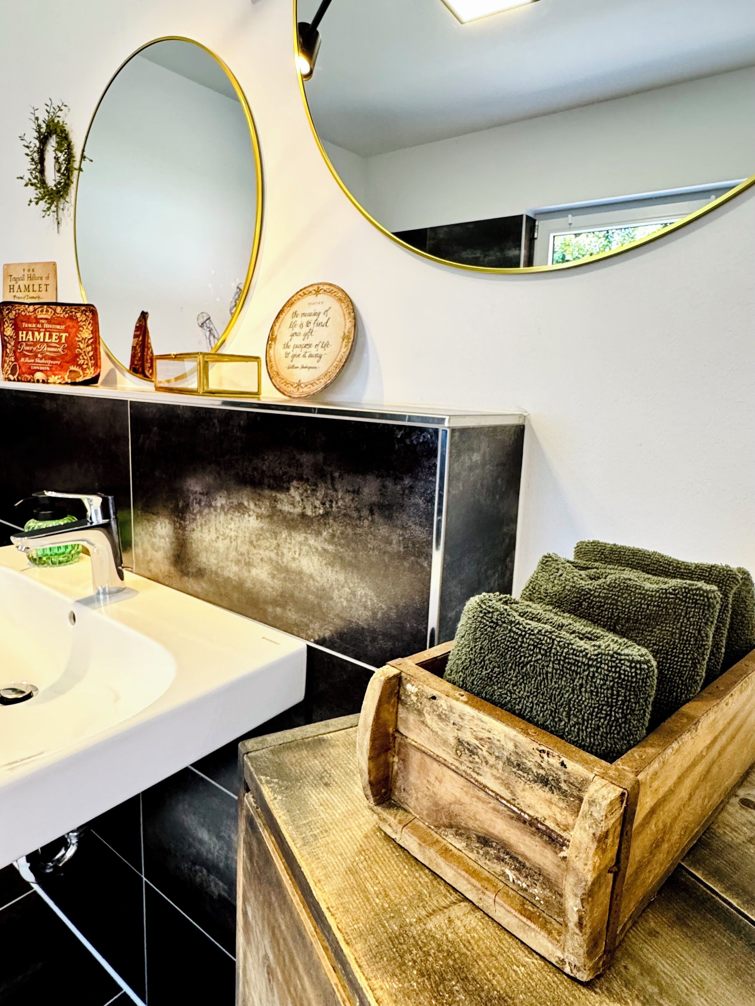 Une salle de bain avec un lavabo blanc, des carreaux noirs, un banc en bois avec des serviettes, un miroir rond et une citation encadrée au mur.