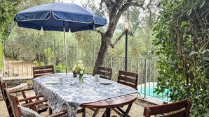 Una terrazza esterna con un tavolo rotondo, sedie, un ombrellone blu e una piscina sullo sfondo.