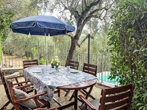 Eine Terrasse im Freien mit einem runden Tisch, Stühlen, einem blauen Sonnenschirm und einem Pool im Hintergrund.