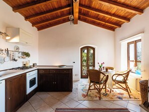 Una cocina con vigas de madera, un comedor con sillas de mimbre y una vista por una puerta.