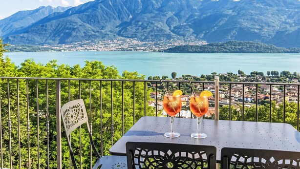Une table avec deux verres d’Aperol Spritz sur un balcon donnant sur un lac et des montagnes.