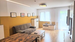 A modern living room with a sofa, a coffee table, and a TV. There are framed pictures on the wall, a window with curtains, and a kitchen area with cabinets and a microwave. - LUFT Apartment in Kritzendorf/Klosterneuburg near Vienna, with parking (Klosterneuburg)