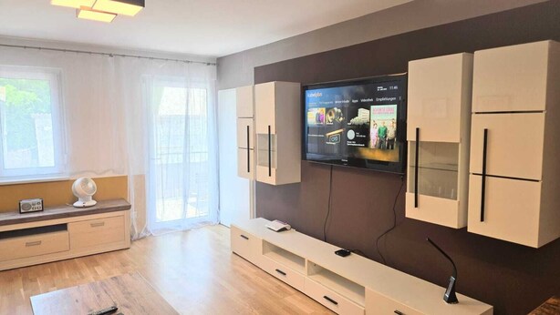A modern living room with a flat-screen TV, built-in cabinets, a wooden floor, and a window with sheer curtains. - LUFT Apartment in Kritzendorf/Klosterneuburg near Vienna, with parking (Klosterneuburg)